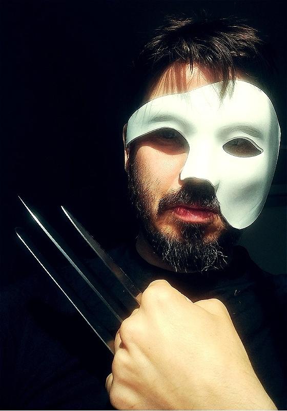 Joz Norris, a white man with brown hair and beard, is looking at the camera wearing a "phantom of the opera" half-face white mask. He is holding his left hand up on his chest with three knives between his fingers, looking like Wolverine's hand.