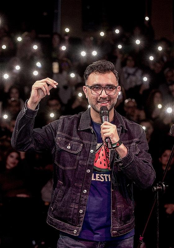 The performer holds a microphone in his left hand with an audience behind him holding their phone flashlights up