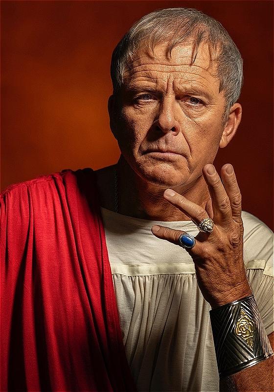 Maxwell Caulfield, a white man with short grey hair, wearing a white Roman shirt with a red robe over his shoulder, is looking intensely at the camera with his hand up, showing two rings and a metal wristband.