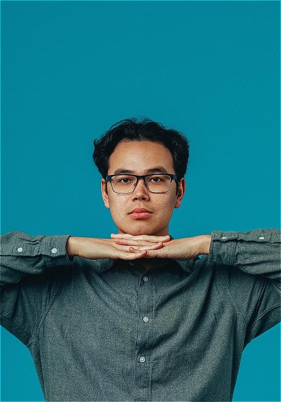 A man wearing glasses in a long sleeve shirt stares at you with his hands interlocked under his chin. 