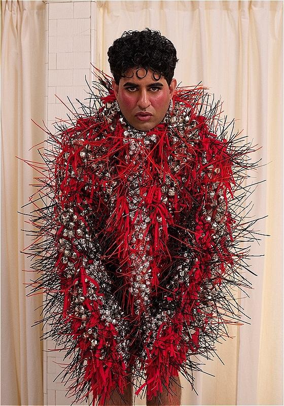 ALOK, a non-binary brown person with short curly black hair and blush on their cheeks, is looking directly at the camera. They are wearing a fashionable coat made of red and black spikes and silver jingle bells, covering their body, neck and hands. The background is made of cream colour curtains.