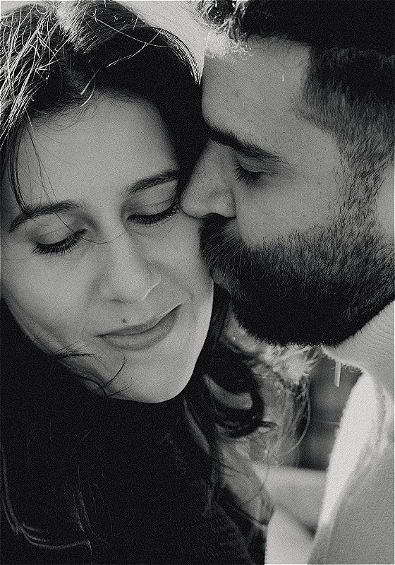 A close-up picture of a brown man with a beard and a brown woman with long black hair. The woman is facing the camera but looking down and smiling as the man is kissing her cheek, eyes closed.