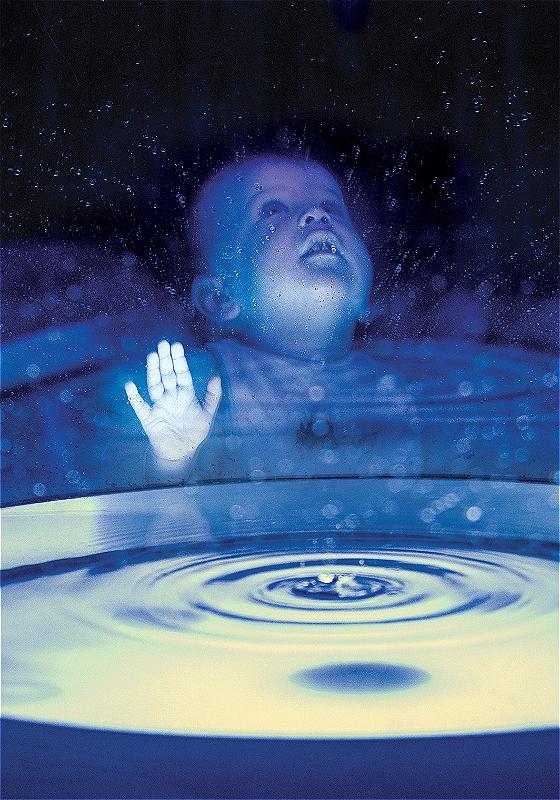 A baby presses their hand against a glass surface, looking up in wonder. In the foreground a pool of water seems to be forming.
