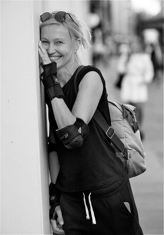 A black and white photo of Ingeborga Dapkunaite, a white woman with blonde hair, sunglasses on her head, a sleeveless black top and wrist and elbow braces. She is looking at the camera smiling, her left hand and side of her face resting on a wall. The background is a blurred street with silhouettes and buildings.