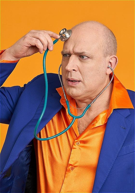 A bald Man dressed in a bright blue and orange suit holds stethoscope to his head. He pulls a concerned expression as he listens to the contents of his brain.
