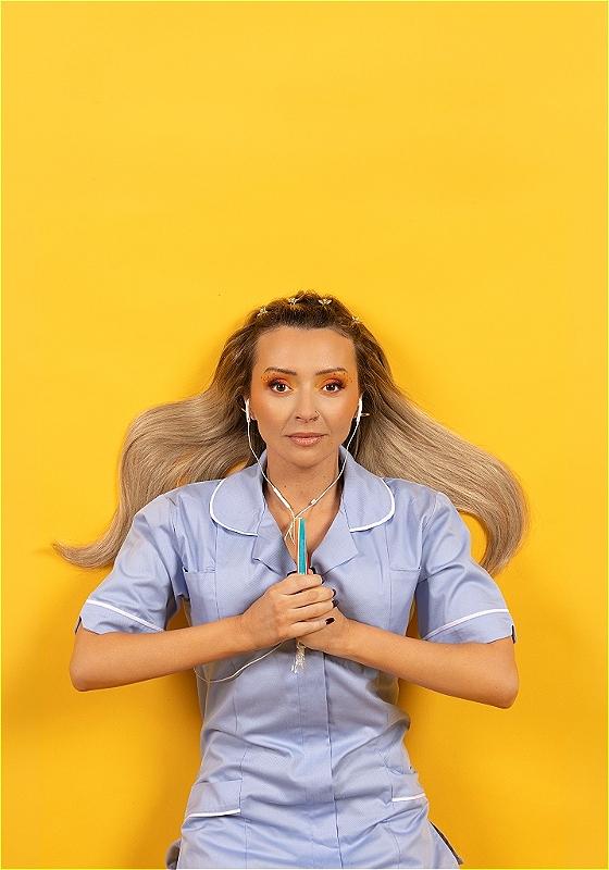 Kimberley is against a bright yellow background. They are wearing a blue nurses dress, with wired earphones in their ears. They are clasping a stick of rock between their hands, in front of their chest.  