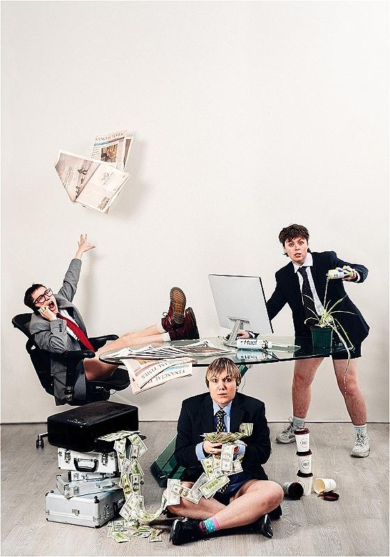 Three people in suit jackets and no trousers sit at a glass table. One shouts loudly on the phone. Another is on a computer whilst watering a plant with an energy drink. The other sits on the floor with loads of money in their hands. 