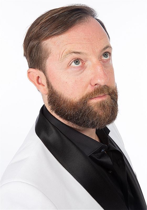 A man in a white suit jacket with black satin lapel, a beard, looks up to the ceiling.