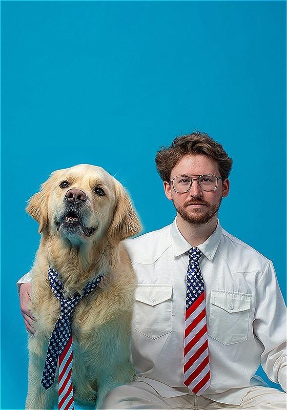 A man and his golden retrierver sit in front of a bright blue background. He wears a shirt and an American flag tie. His dog also wears an american flag tie.  