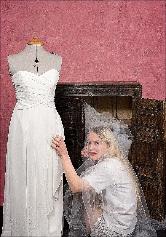 A woman crouches to the right of a mannequin which has a wedding dress on it. She is wearing the veil which covers her whole body. She looks scared.