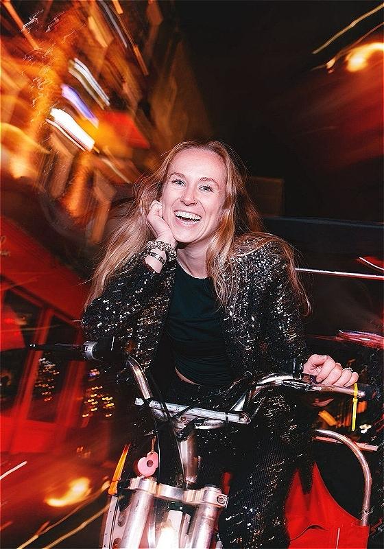A woman with long blonde hair rides through the streets of London at night on a tuk tuk. 
