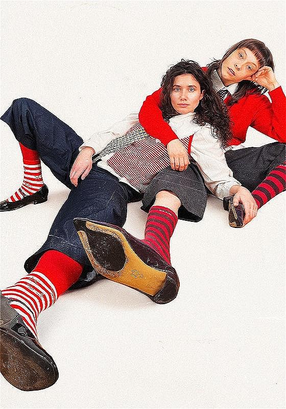 Two female presenting people sit on the floor in a relaxed manner. One lies across the other. They are wearing stripey socks, blue/black jorts, white shirts and ties. It looks very preppy. 