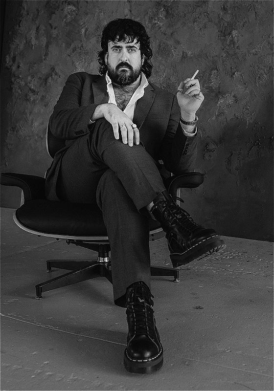 A black and white photo of Zachary Ruane, a white man with a black mullet and beard, wearing a grey suit, white shirt and chunky black boots. He is seriously looking at the camera, holding a cigarette in his left hand and sitting in an office chair. 