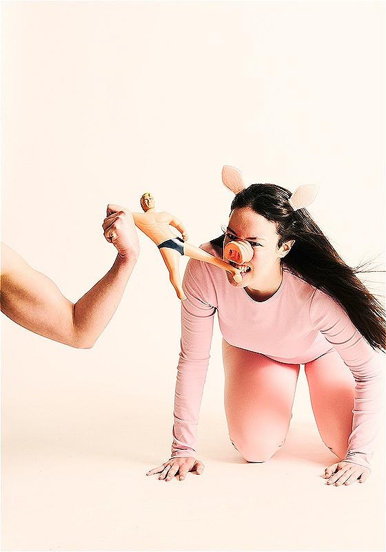 A woman dressed as a pig holds a ken doll in her mouth, whilst a man flexing his arm holds the other side of the doll. 