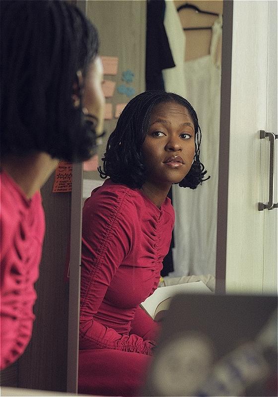 Ayoade Bamgboye, a black woman with shoulder length locks and a red long-sleeve top, is looking at herself in the mirror of a bedroom wardrobe. In the background we can see post-it notes, a white dress and her laptop.