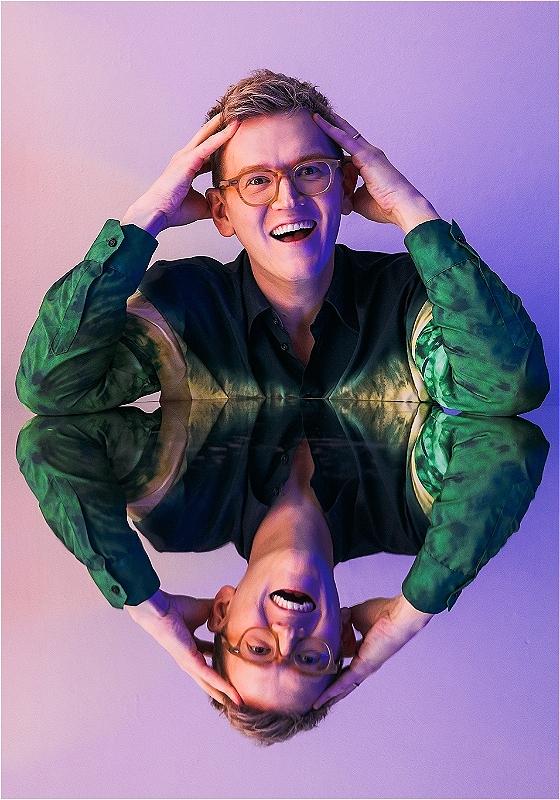 Chris Parker, a white man with round glasses and short blond hair, is smiling at the camera with his hands in his hair. He is resting his elbows on a mirrored surface, making the lower half of the photo his upside-down reflection.