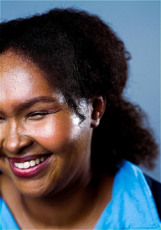 The performer Maryam Garad, a Black woman with black afro hair and dark pink lipstick, is smiling with her eyes closed as if she's mid-laughter..