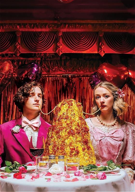 Two individuals dressed entirely in pink sit at a table covered in roses and eat from a large pile of spaghetti.