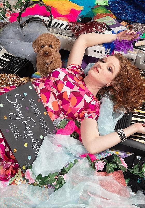An individual lies amongst a pile of roses, keyboards and sequins. A chalk board rests against her lap with the words 'Please Submit Song Requests Here' written on it.