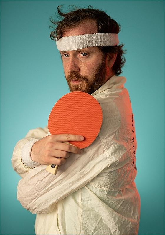 A man in a white shell jacket, holding an orange ping pong bat and wearing a white headband looks at us with intrigue. 