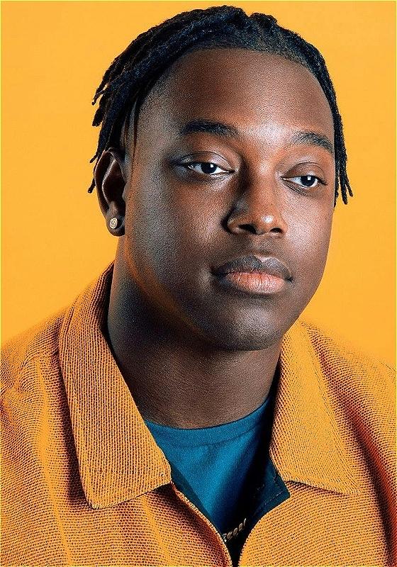 Ify, wearing an orange shirt against an orange background, looks blankly off. 