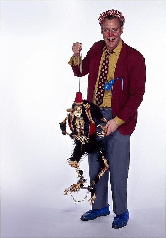 Jeremy Lion, a white man with short ginger hair, wearing a red flat cap, a yellow shirt with a red jacket and a red polka dot tie, is smiling at the camera holding a monkey puppet on a string, over a white background.
