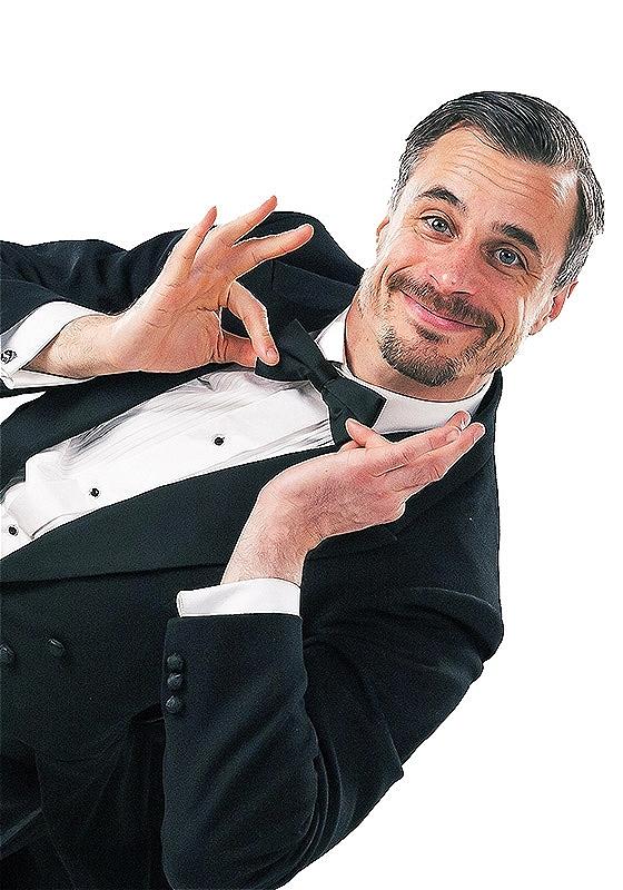 A man in a black morning suit and bow tie, with slicked back hair, twizzles his bowtie and leans on one foot to the right, at a dangerous angle.
