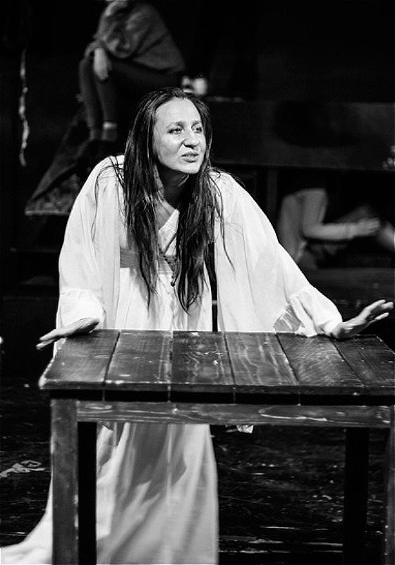 A black and white image of an individual with long black hair mid speech. Their palms are pressed against a wooden table and they are dressed in a long white nightgown.