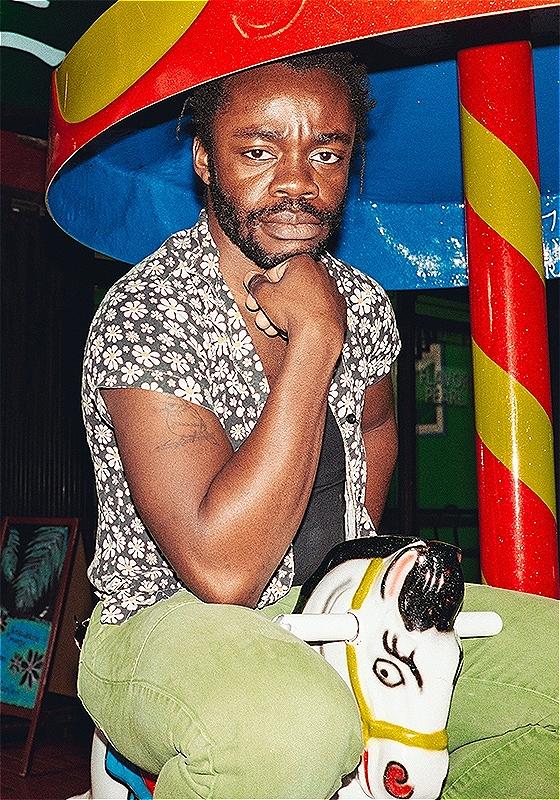 Demi Adejuyigbe, a Black person with short afro hair and a beard, wearing a flowery shirt and green trousers, is sitting on a small carousel horse, looking seriously at the camera. Over him is the umbrella shaped top of the carousel and the background is a shopping centre.