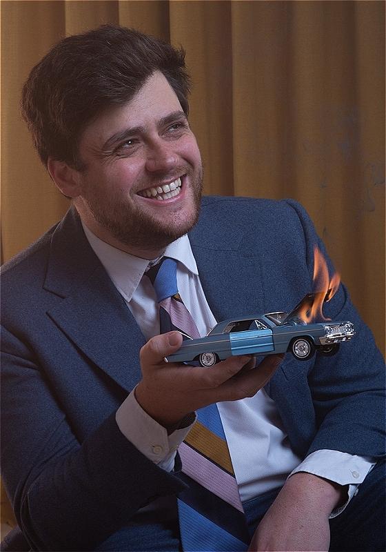 Bróccán Tyzack-Carlin, a man with brown hair and stubble, is wearing a blue suit and white shirt with a colourful tie. He is smiling looking to the side and holding a miniature car with the hood on fire, over a brown curtain background.