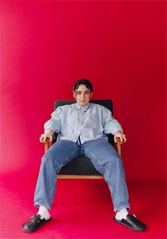 An individual is slumped on a chair and staring expressionless at the camera. The floor and wall are painted a vivid red.