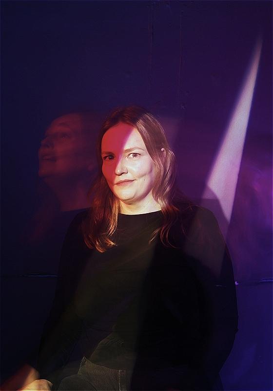 Kate is looking into the camera. A beam of light illuminates half of her face. 