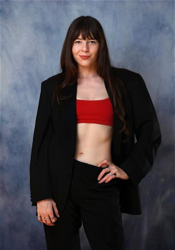 Chelsea, wearing a red sports bra over a black blazer, smiles at the camera before a grey background.