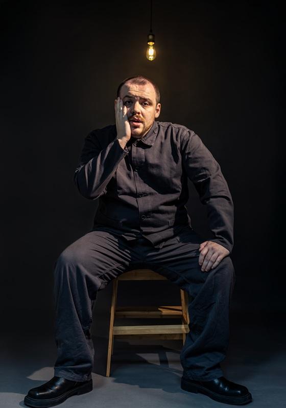 A person is in a dark empty room, beneath a start bare lightbulb. They're wearing all-black and they look worried, holding a hand up to their bearded face. 
