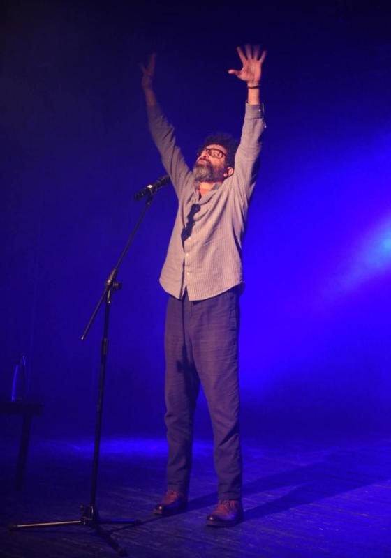 A performer lifts their arms up as they speak into a microphone. The stage they are on is submerged in a deep blue lighting.