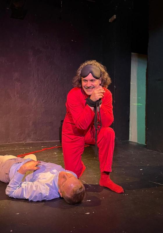 A man wearing an all red outfit crouches next to a body. His hands are bound but he smiles out into the audience.