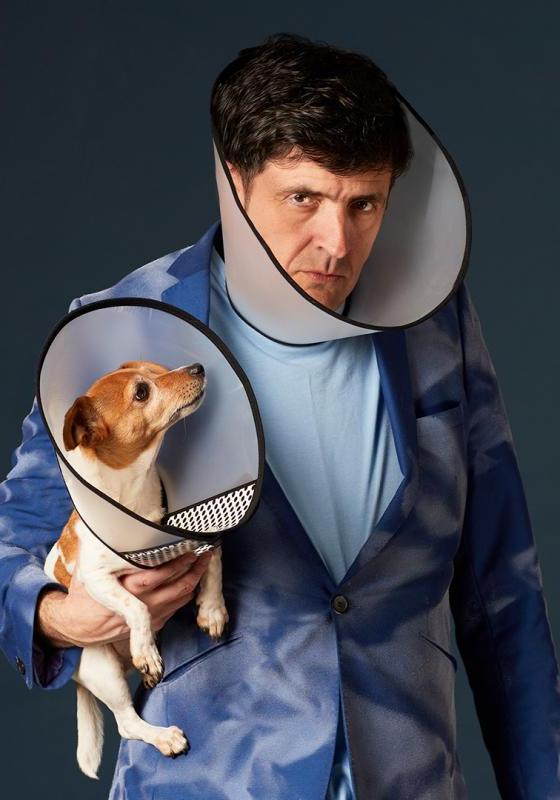 Spencer Jones wears a dog cone and blue suit and holds a small dog also wearing a cone. 