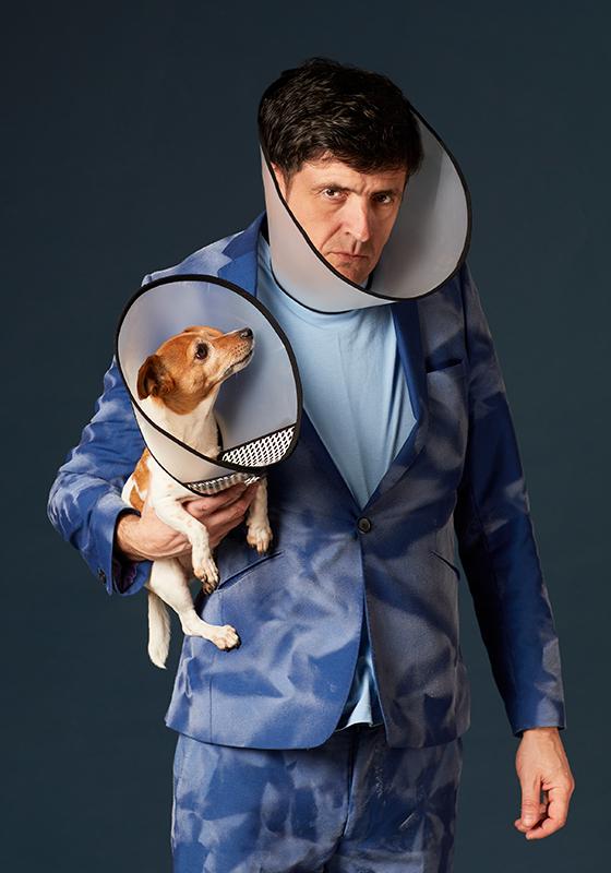 Spencer is looking disgruntled, wearing a blue blazer. He's holding a dog with a cone around it's neck. The dog is looking up at Spencer, who is also wearing a cone around his neck. 