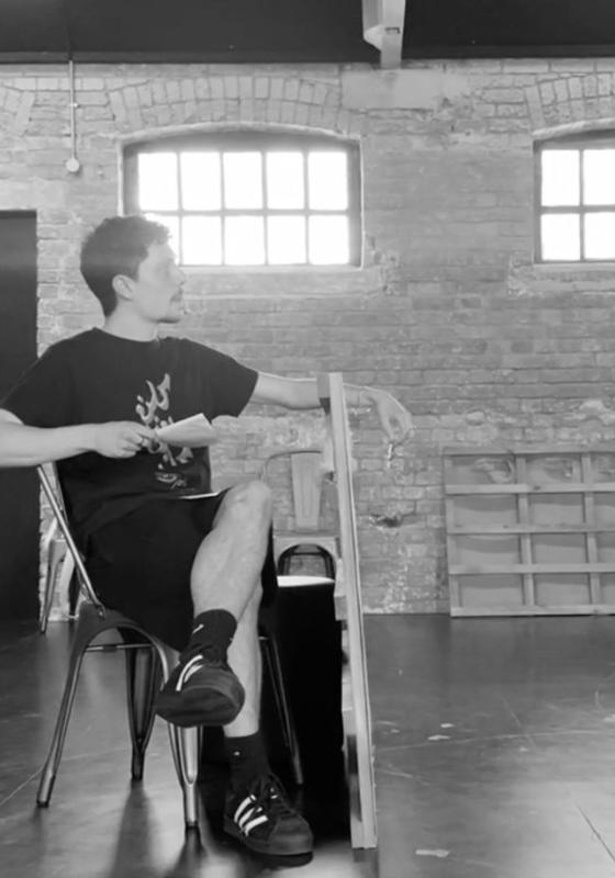 A black and white image of two people seemingly rehearsing. One is sat down while the other is stood. Both of the performers are holding a piece of paper.