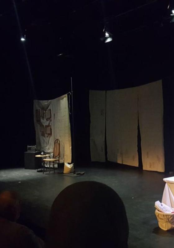 An image of a stage with sheets hung across the back. Centre stage is a table with a sewing machine. A woman dressed in a headscarf is crouched over the table seemingly sewing.