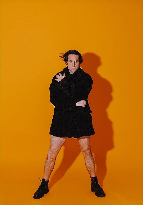 Luke stands against a bright orange backdrop, wearing just a black fluffy coat and some black boots. 