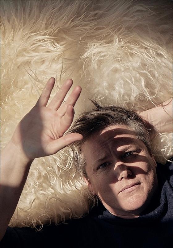 Hannah Gadsby lies on a bed of white fur. They look up at the camera and block the sunlight with their hand.
