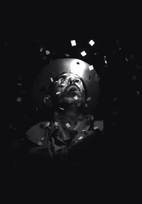 A man in darkness looks up at a bright light from above. Confetti falls around him. 