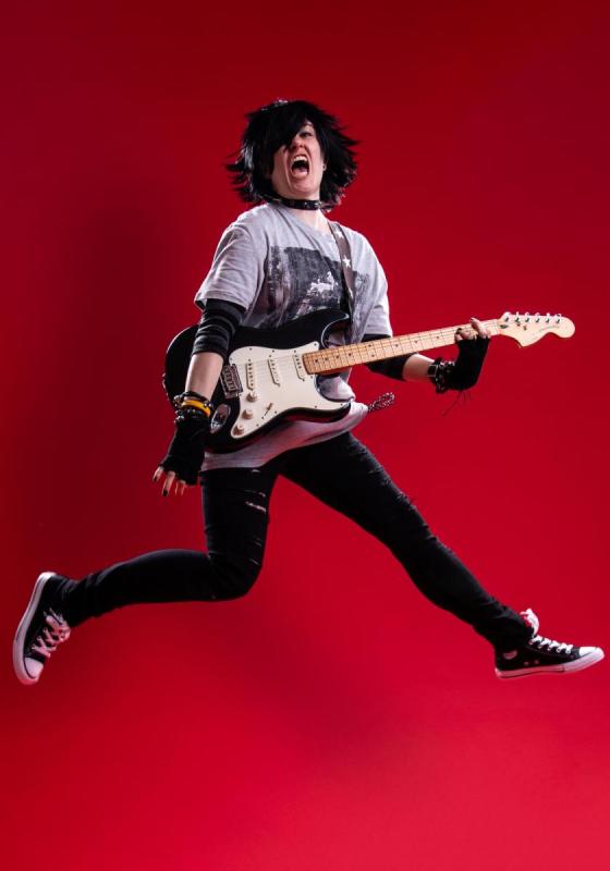 The performer in a rock-emo outfit holds a guitar, jumping in midair in front of a red background