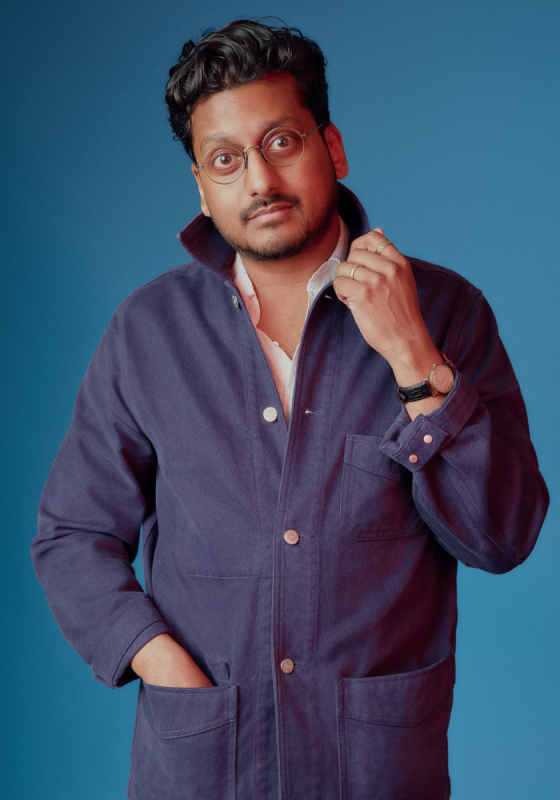 Ahir satnds in front of a bright blue back drop. They are looking calmly into the camera as they fiddle with the collar of their blue boiler suit.