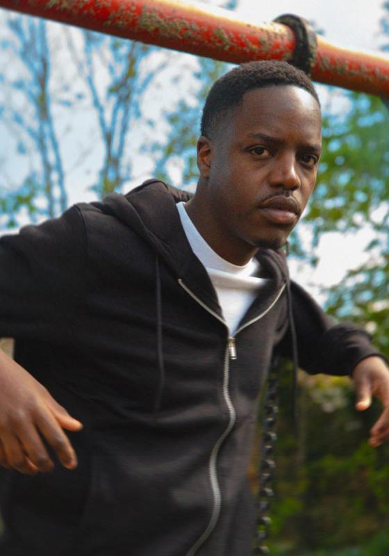Performer stands in a park during the day, wearing a black hoodie