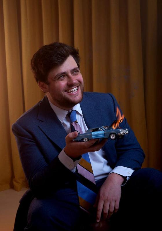 Bróccán Tyzack-Carlin, a man with brown hair and stubble, is wearing a blue suit and white shirt with a colourful tie. He is smiling looking to the side and holding a miniature car with the hood on fire, over a brown curtain background.