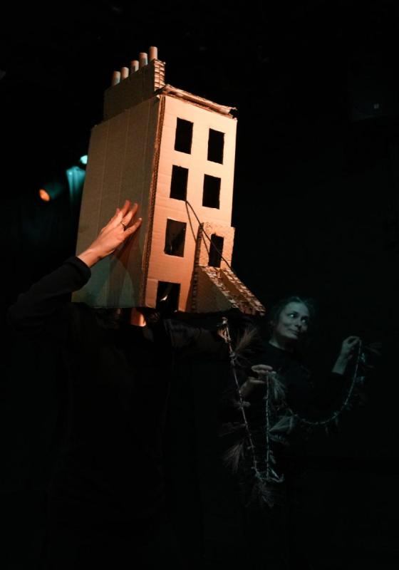 Performers in the dark hold up a brightly lit cardboard townhouse