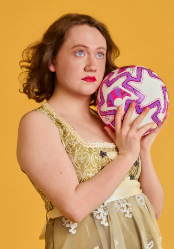 Dee stands in front of a yellow back drop. They are holding white, pink and purple football and gazing into the distance.