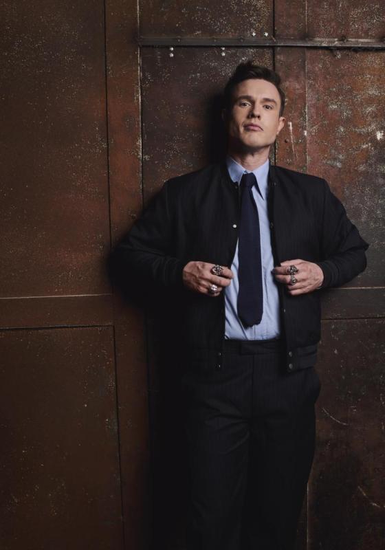 Ed Gamble leans against a wooden wall. He is smartly dressed in a blue shirt and black tie, and looks confidently into the camera.